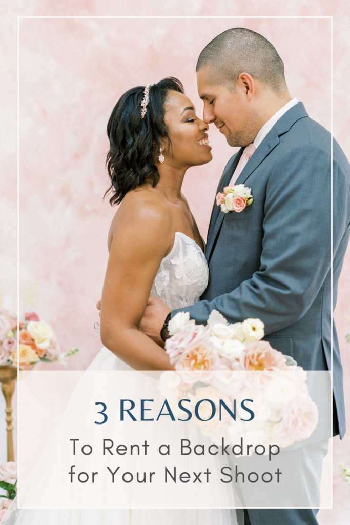 A bride and groom posing facing each other in front of a blush hand painted backdrop. The bride is a black woman with short hair and she is holding a bouquet of white, blush and light orange flowers. The groom is hispanic and wearing a grey suit.
